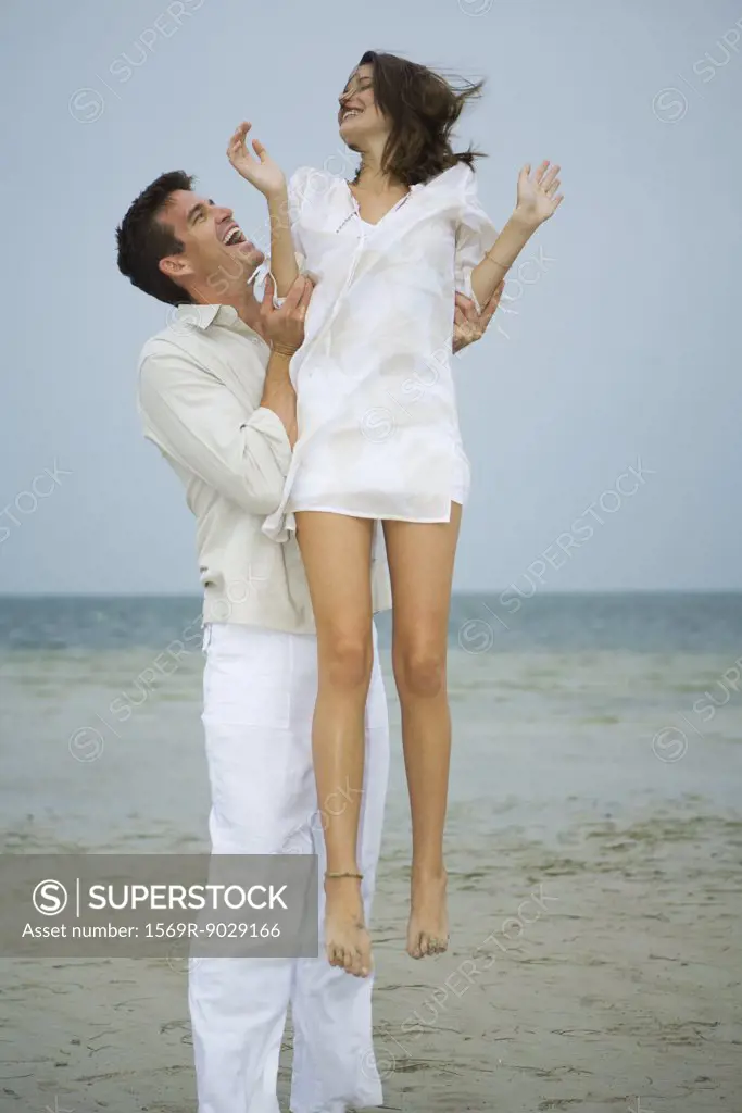 On beach, man holding young female companion up in air, full length