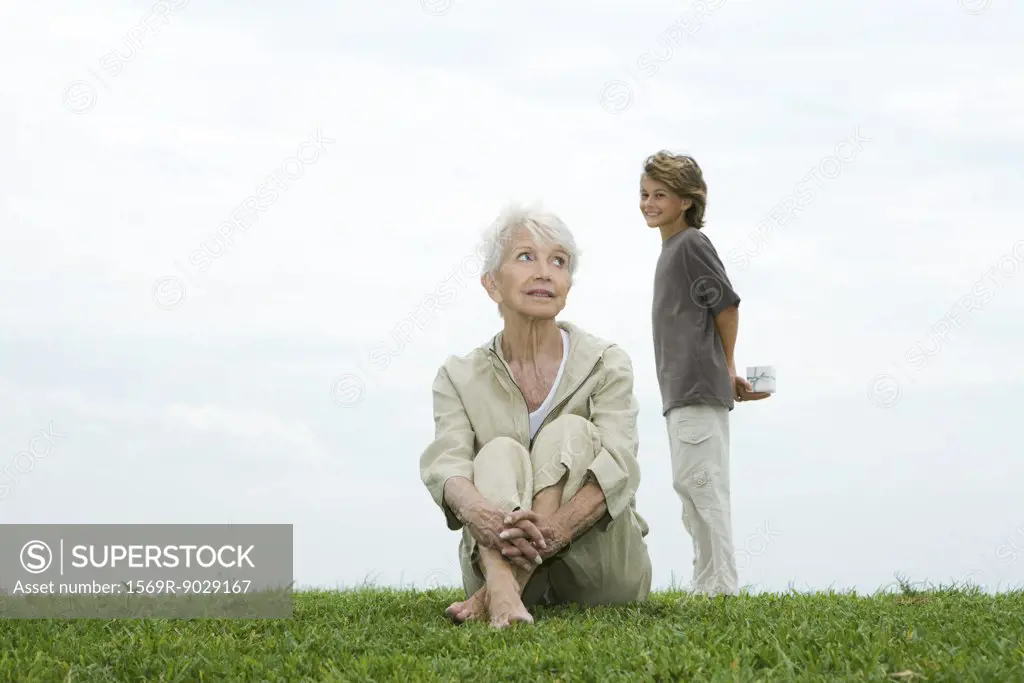 Senior woman sitting on grass, grandson standing behind her, holding gift, smiling at camera, full length