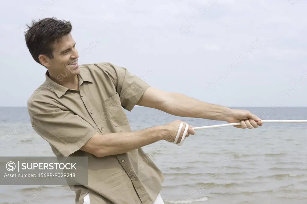 Man pulling rope, smiling, ocean in background, waist up - SuperStock