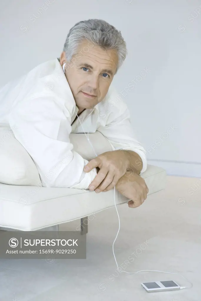 Mature man lying on stomach on chaise longue, listening to MP3 player, looking at camera