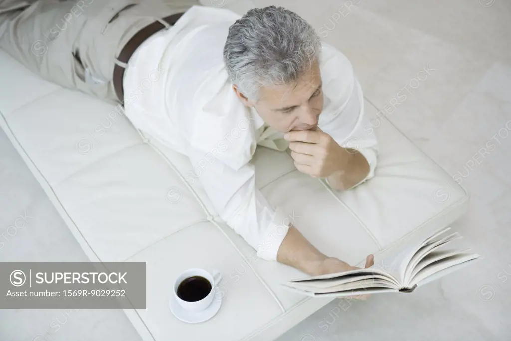 Mature man lying on stomach on chaise longue, reading book, cup of coffee nearby, high angle view