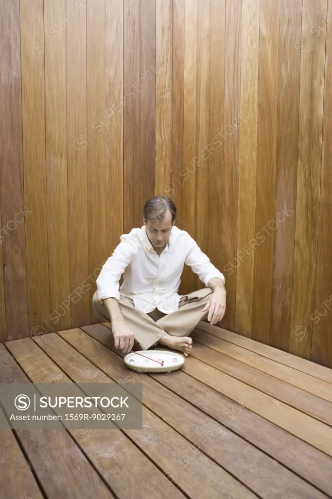 Man sitting with legs crossed on wooden floor, looking down at miniature rock garden, full length