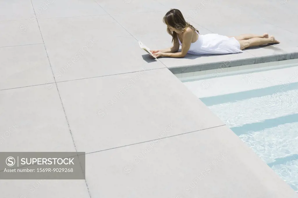 Woman in sundress lying by edge of pool, reading, full length