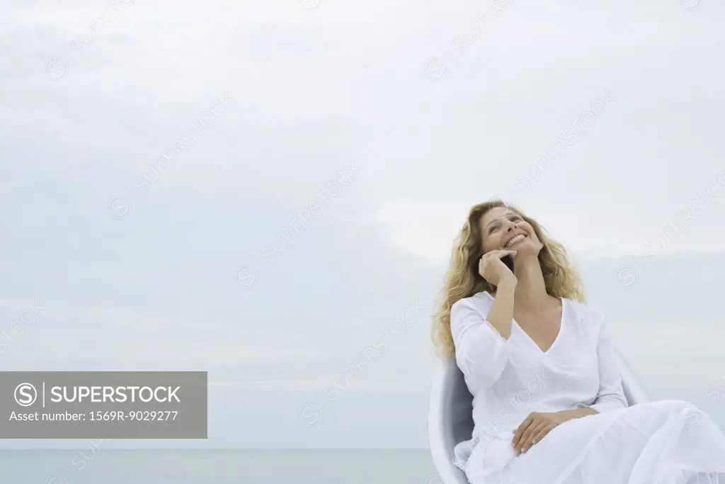 Blonde woman sitting in chair using cell phone, head back and smiling, ocean in background