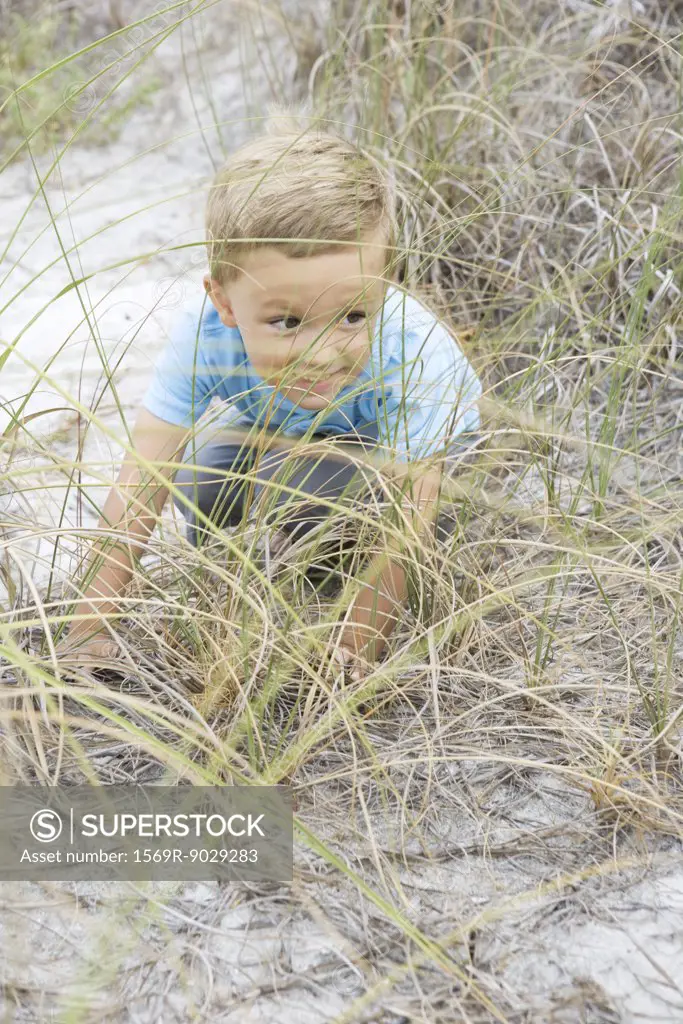 Boy crouching in dune grass, looking away mischievously, full length, high angle view