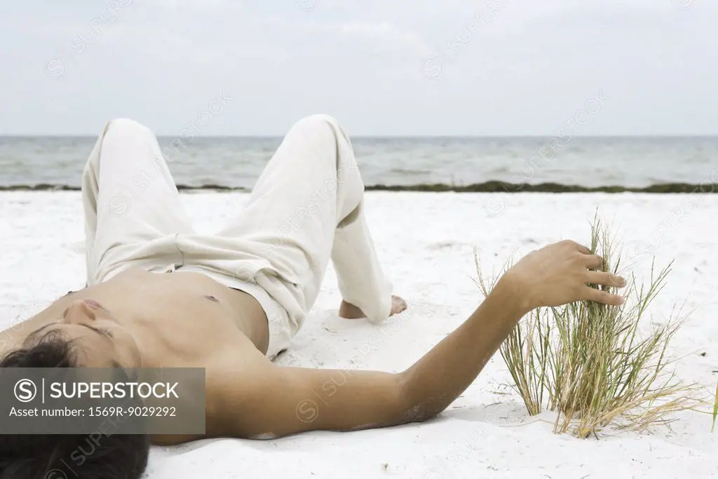Young man lying on back on sand, touching dune grass, ocean in background