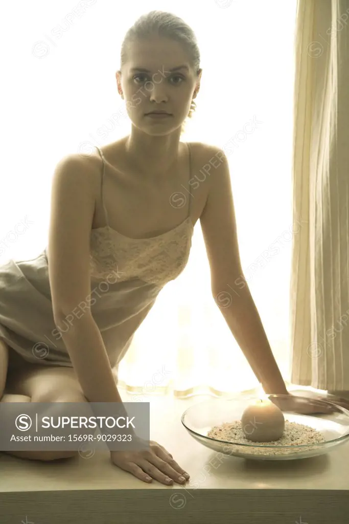 Teenage girl in slip sitting next to candle, looking at camera, backlit