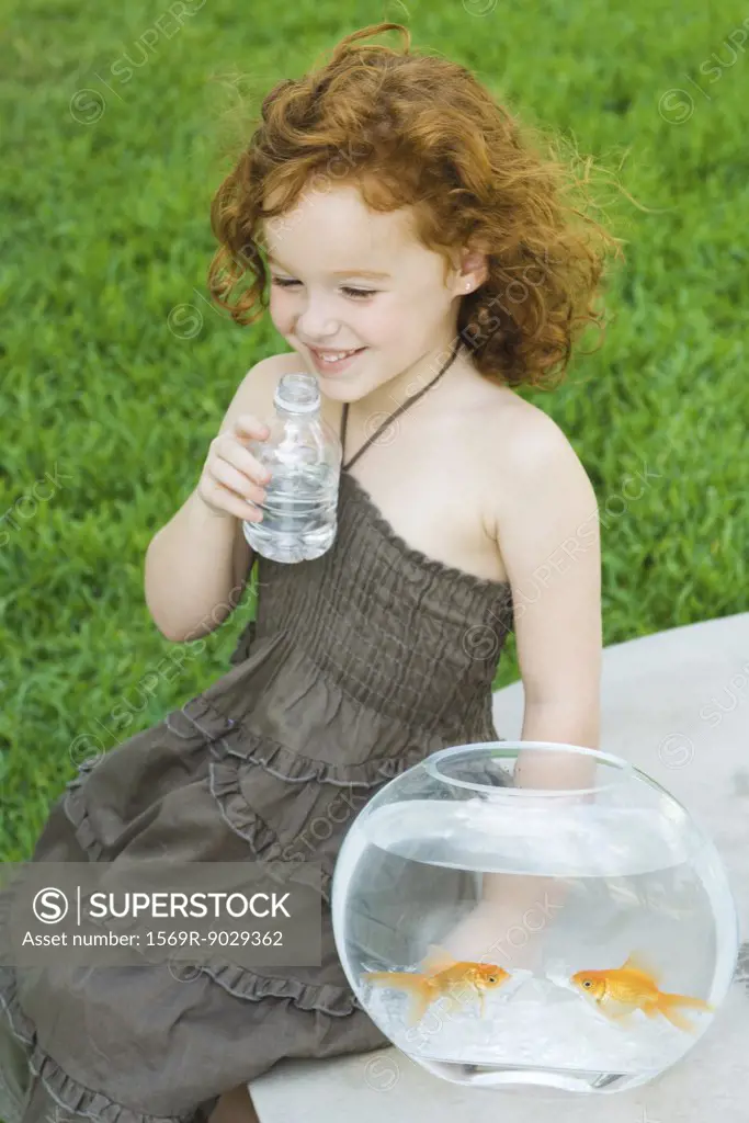 Girl sitting next to goldfish bowl, drinking water