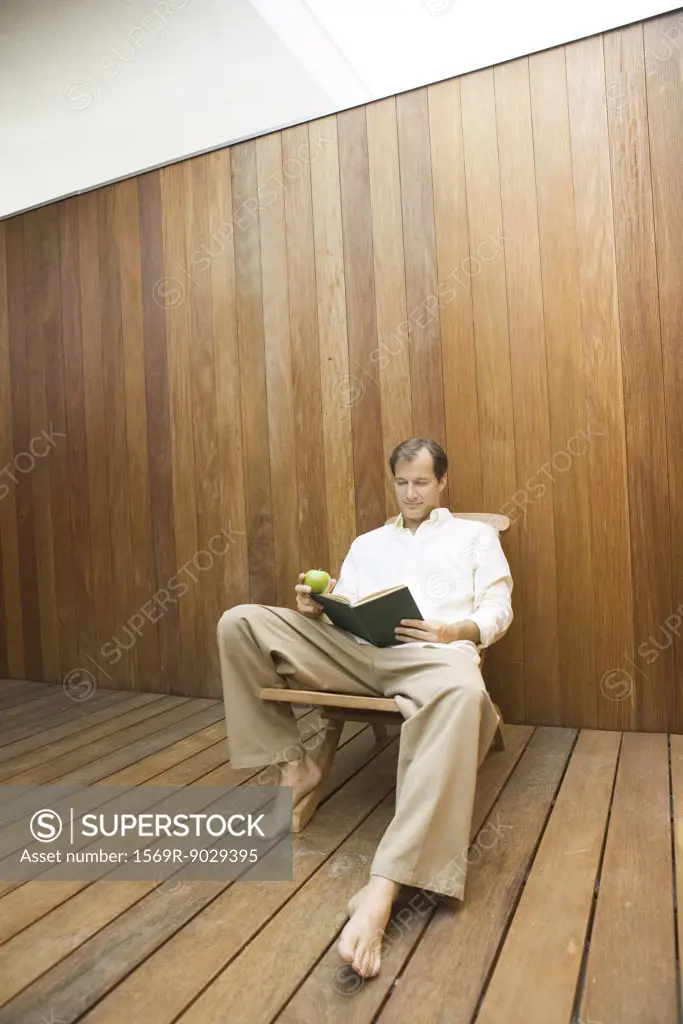 Man sitting in lounge chair reading book, apple in hand