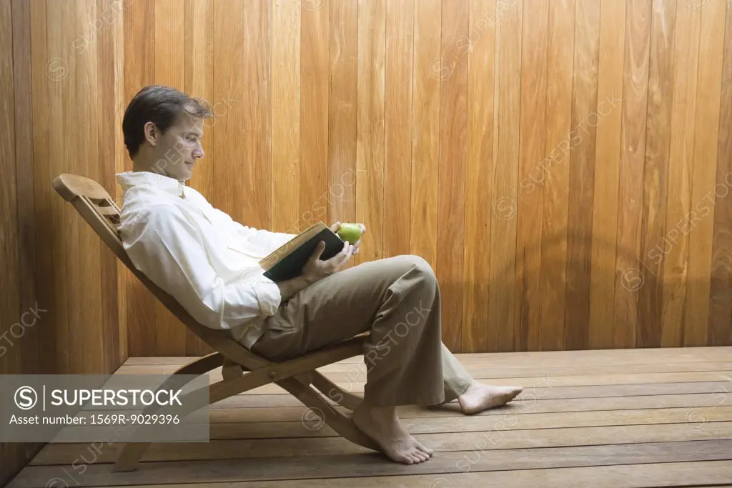 Man sitting in lounge chair reading book, apple in hand, side view