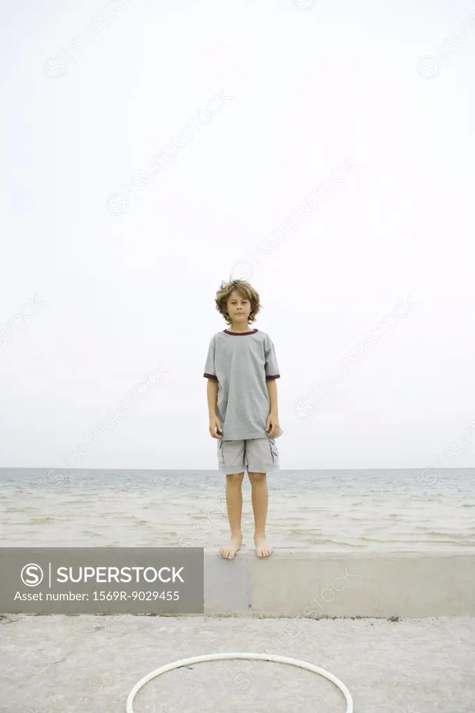 Boy standing on low wall at the beach, looking at camera