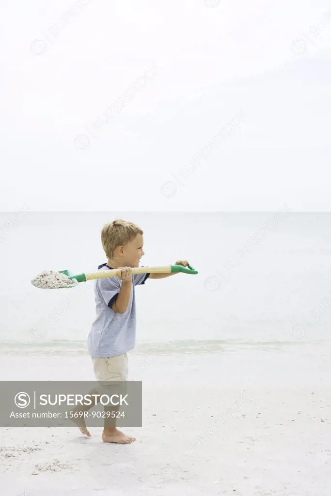 Boy walking on the beach, carrying shovel full of sand