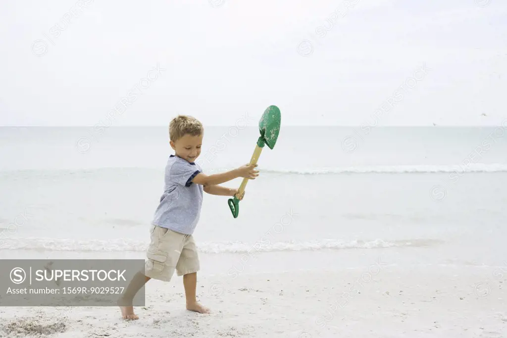 Young boy standing on beach, holding up shovel, looking down