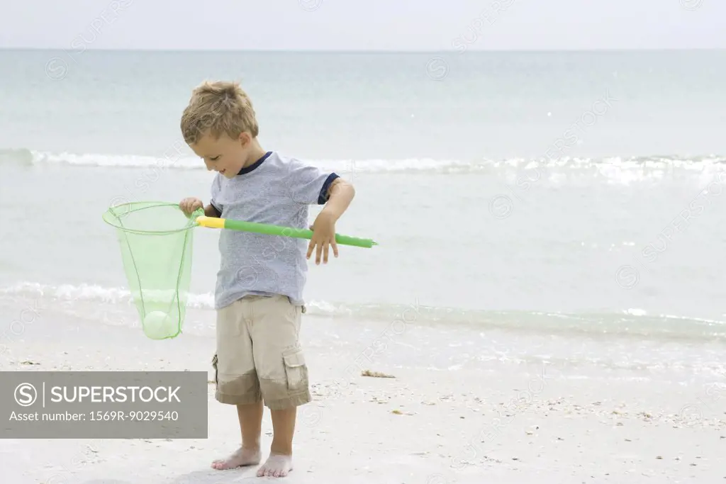 Young boy standing at the beach, holding ball in net, looking down