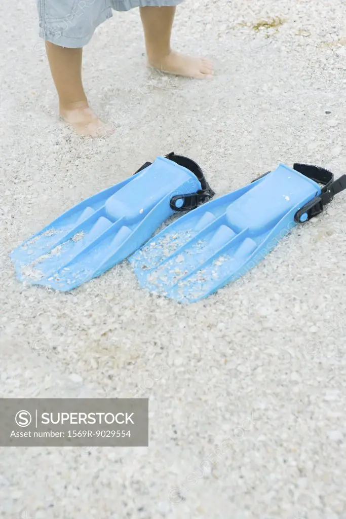 Child standing next to flippers on sand, cropped view