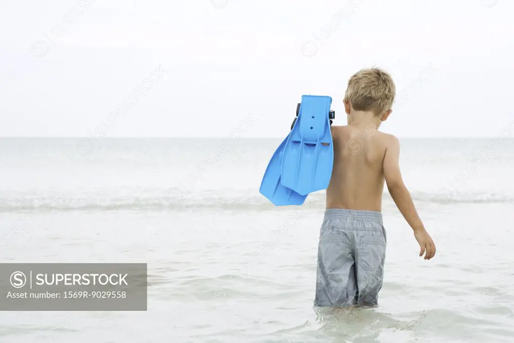 Young boy wading in ocean, carrying flippers, rear view