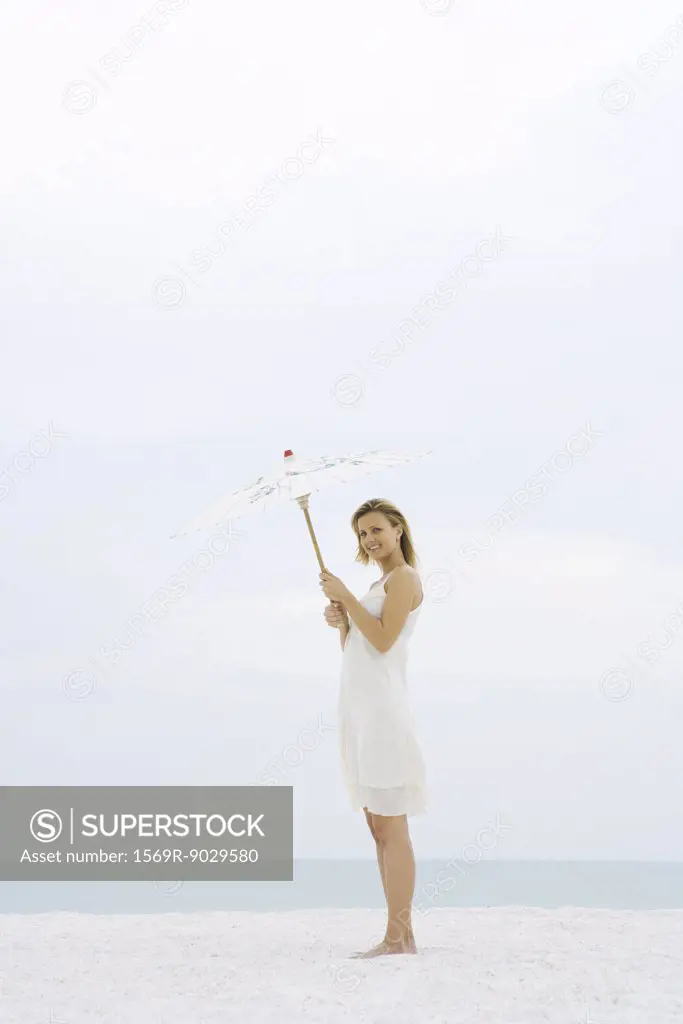 Woman in sundress standing at the beach, holding up parasol, smiling at camera