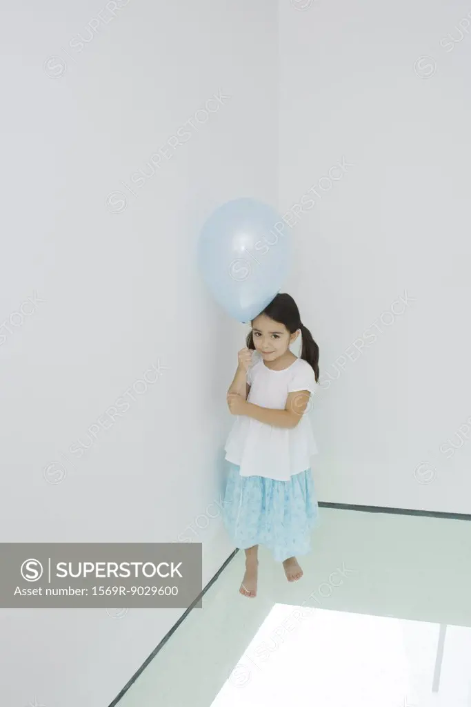Girl standing in corner holding balloon, looking up at camera