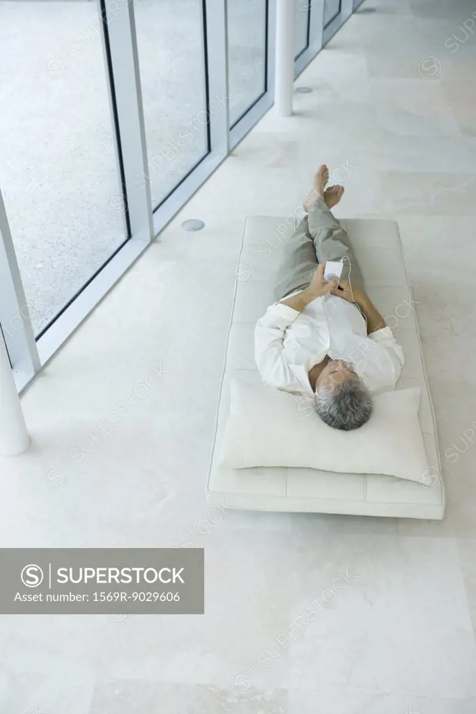 Mature man lying on back on chaise longue, listening to mp3 player, high angle view