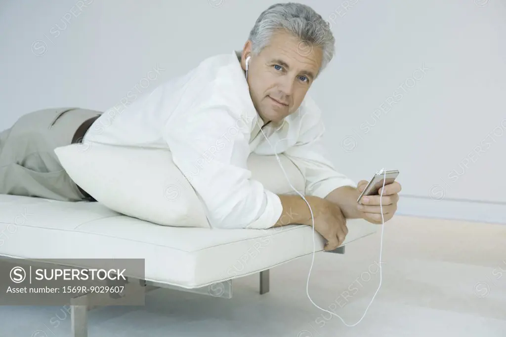 Mature man lying on stomach on chaise longue, listening to mp3 player, looking at camera