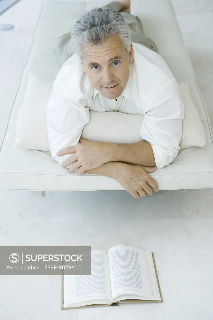 Mature man lying on stomach on chaise longue in front of book, looking up at camera