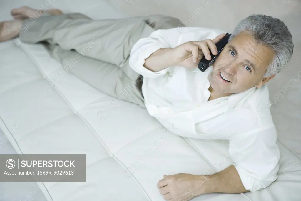 Mature man lying on chaise longue using cordless phone, looking up at camera