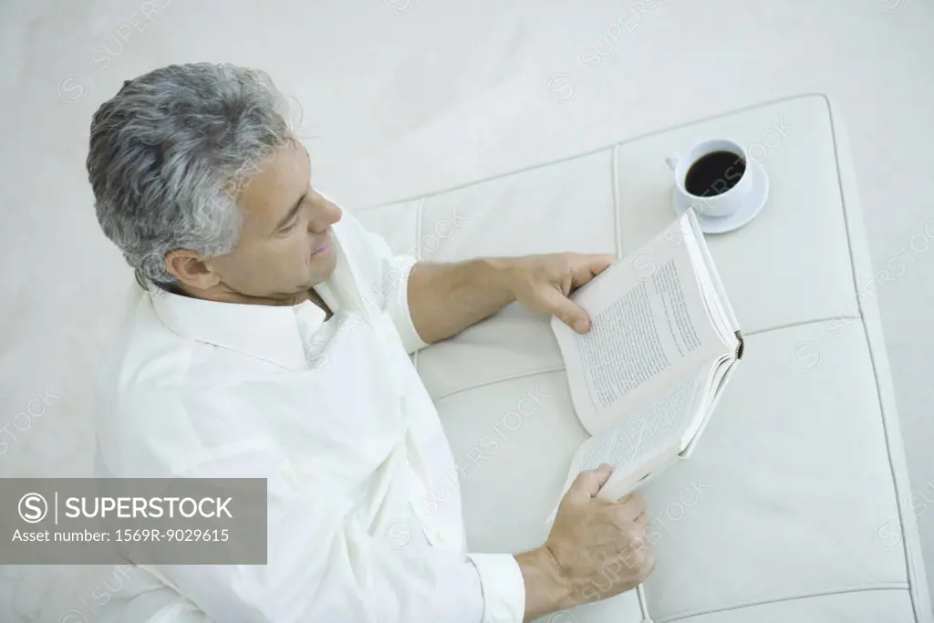 Mature man lying on side next to coffee cup, reading book, high angle view