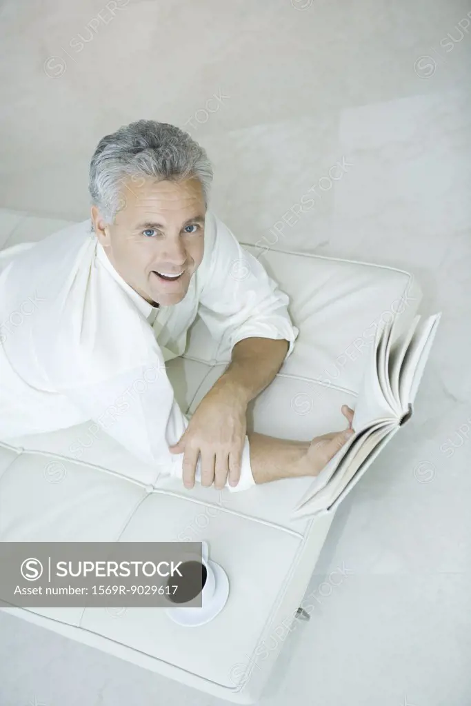 Mature man lying on chaise longue holding book, looking up at camera, high angle view