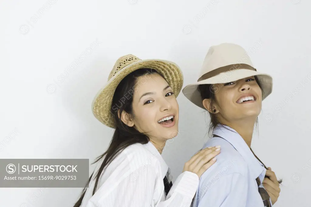 Two young female friends wearing hats and ties, smiling at camera, one placing hand on the other's shoulder