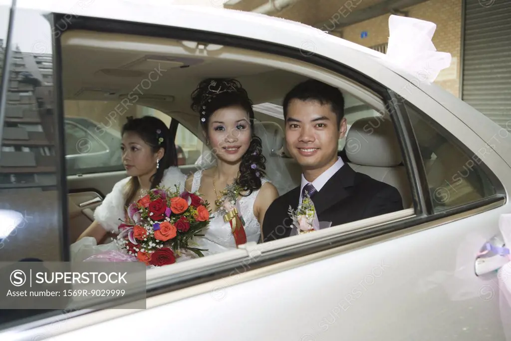 Bride and groom in car with bridesmaid, smiling at camera