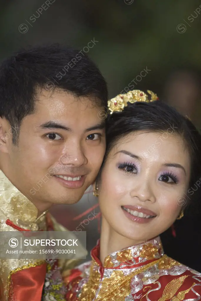 Newlyweds in traditional Chinese clothing, cheek to cheek, smiling at camera