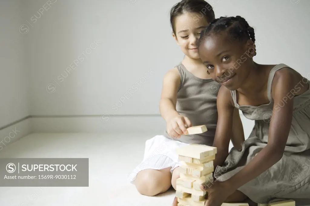 Two girls crouching, building tower with blocks together, one smiling at camera