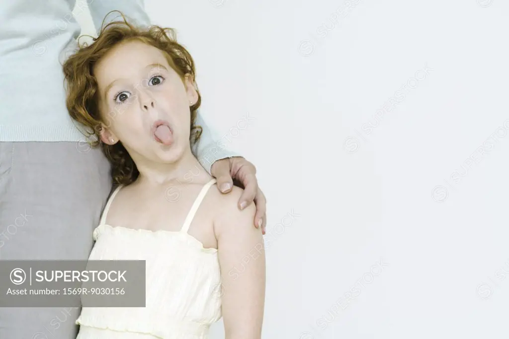 Little girl holding onto mother's leg, sticking tongue out at camera