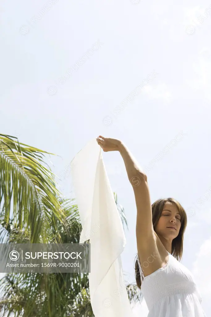 Woman standing in sun in tropical setting, holding up wrap, eyes closed, low angle view