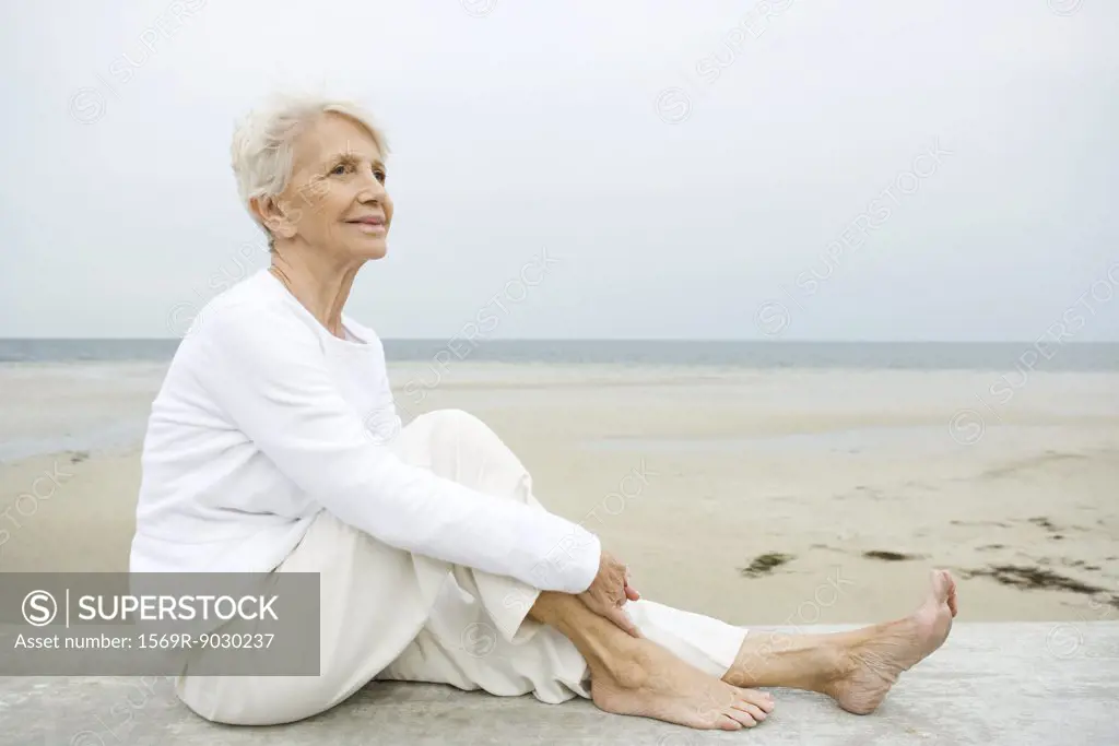 Senior woman sitting on low wall, beach in background, full length