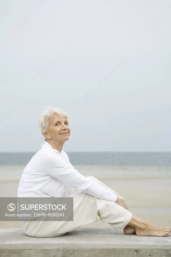 Barefoot senior woman sitting on low wall, smiling, beach in background, full length