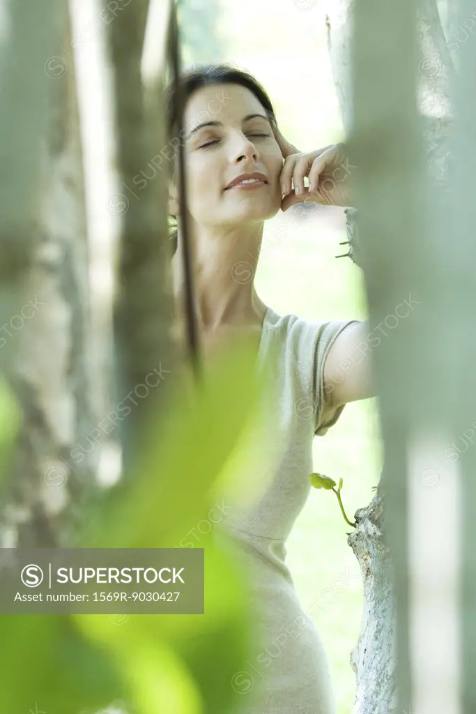 Woman leaning against tree, smiling, eyes closed, view through foliage