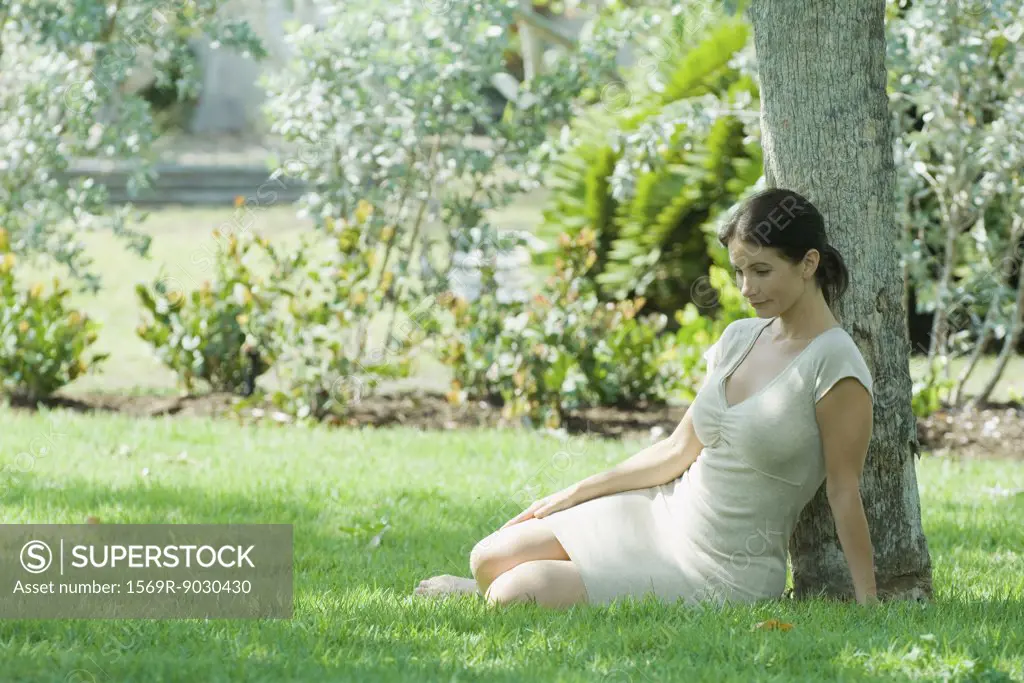 Woman sitting on the ground under tree, smiling, looking down