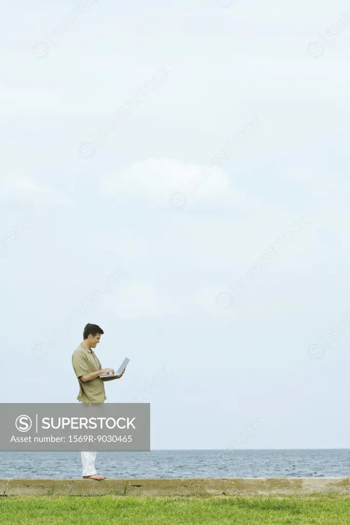 Man standing on low wall using laptop, body of water in background, full length