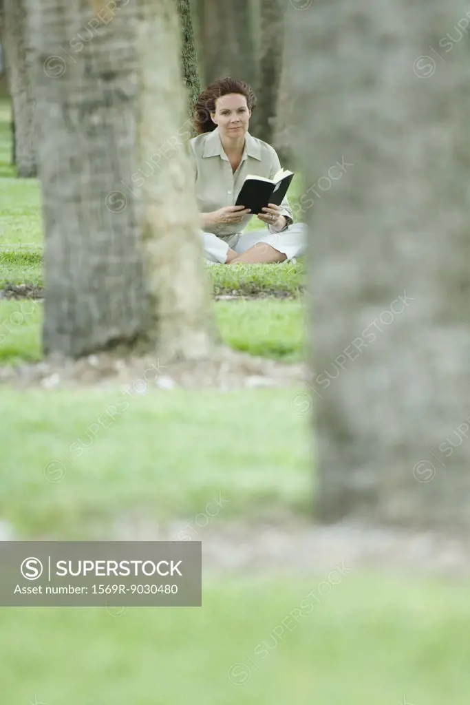 Woman sitting on ground reading book, surrounded by trees