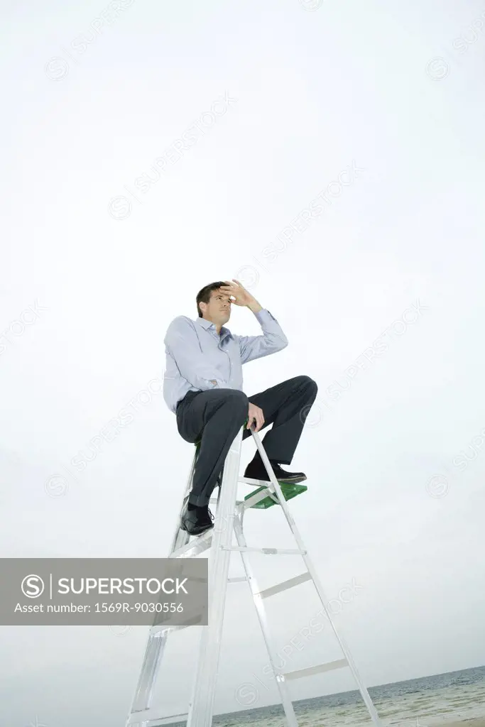 Man sitting on top of ladder, shading eyes, looking away