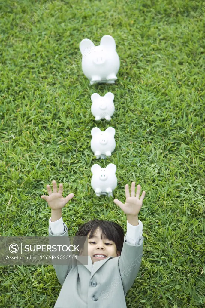 Little boy in full suit lying on grass, line of piggy banks above head