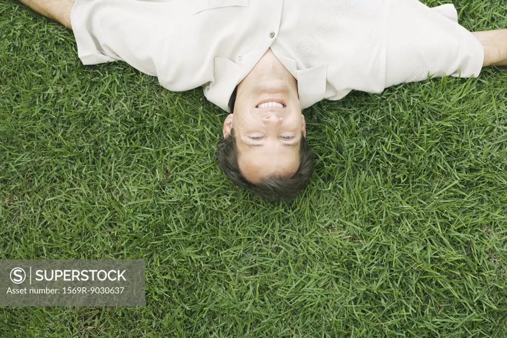 Man lying upside down in grass, smiling at camera, cropped high angle view