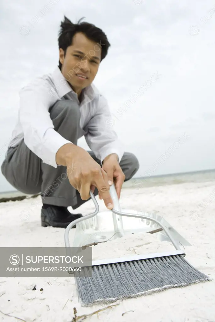 Man crouching on beach, sweeping sand into dustpan, looking at camera, low angle view