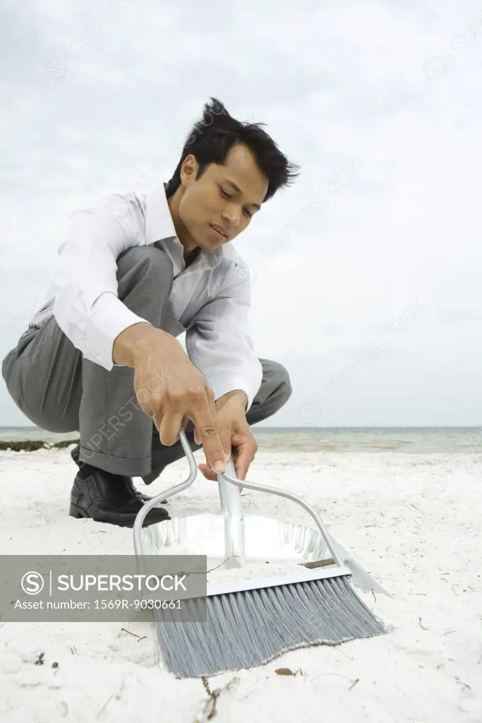 Man crouching on beach, sweeping sand into dustpan, low angle view
