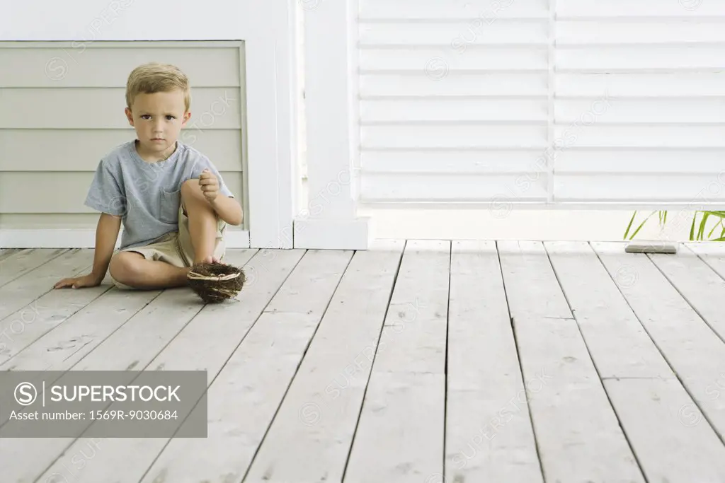 Boy sitting on the ground with bird's nest under his foot, frowning at camera