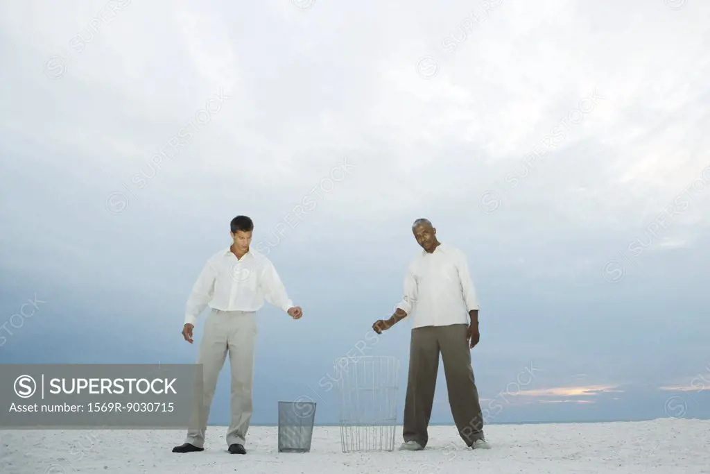 Two men at the beach holding their hands over garbage cans, one looking at camera