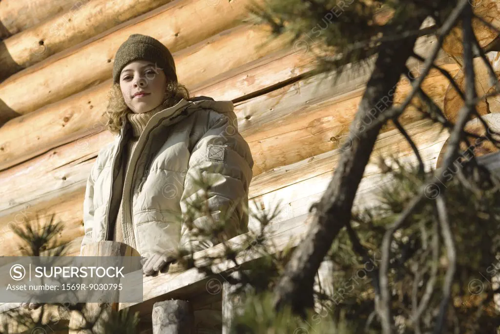 Teen girl standing on deck of log cabin, evergreen in foreground
