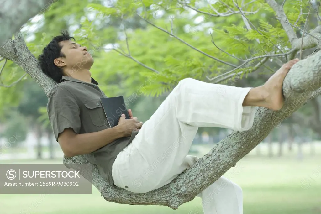 Man resting on tree branch with eyes closed, holding book