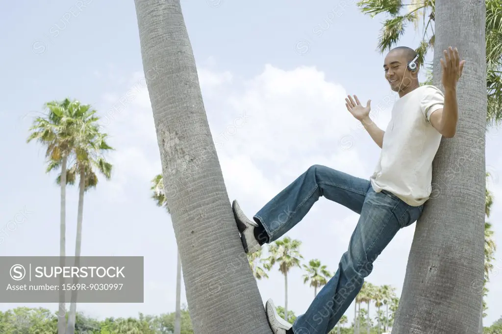 Man leaning against tree trunk listening to headphones, arms out, hands raised